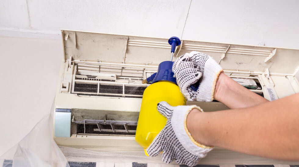 A technician wearing gloves uses a spray bottle to clean the inside of an air conditioning unit.