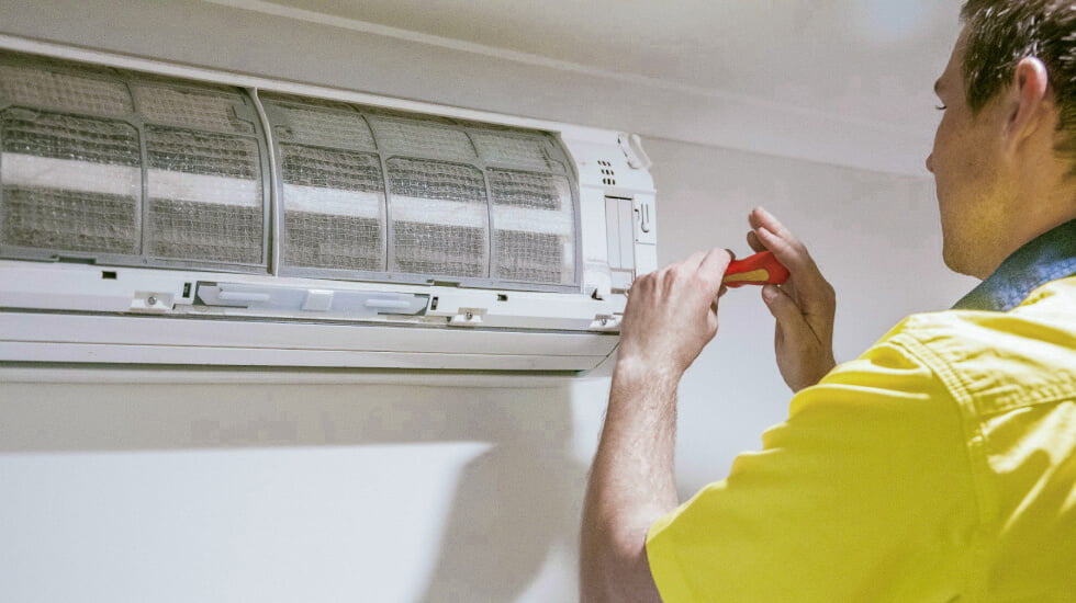A Tenmen Electrical technician in a yellow shirt using a screwdriver to service an indoor air conditioning unit.