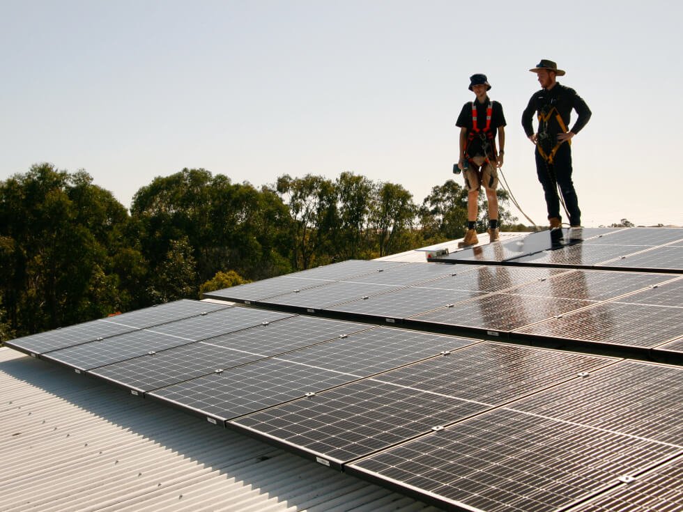 Two workers in safety harnesses stand on a roof inspecting an array of solar panels with trees visible in the background.