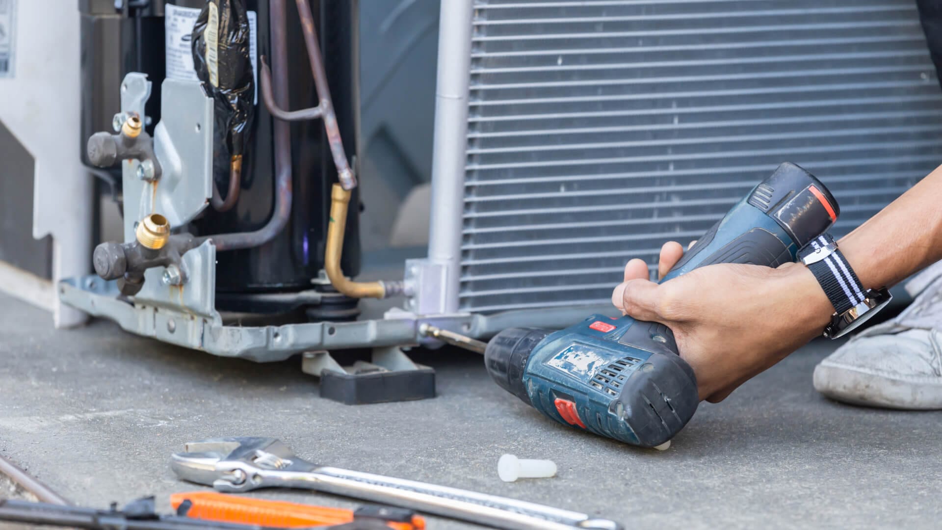 Person using a power drill on HVAC equipment, with various tools nearby on the concrete surface.