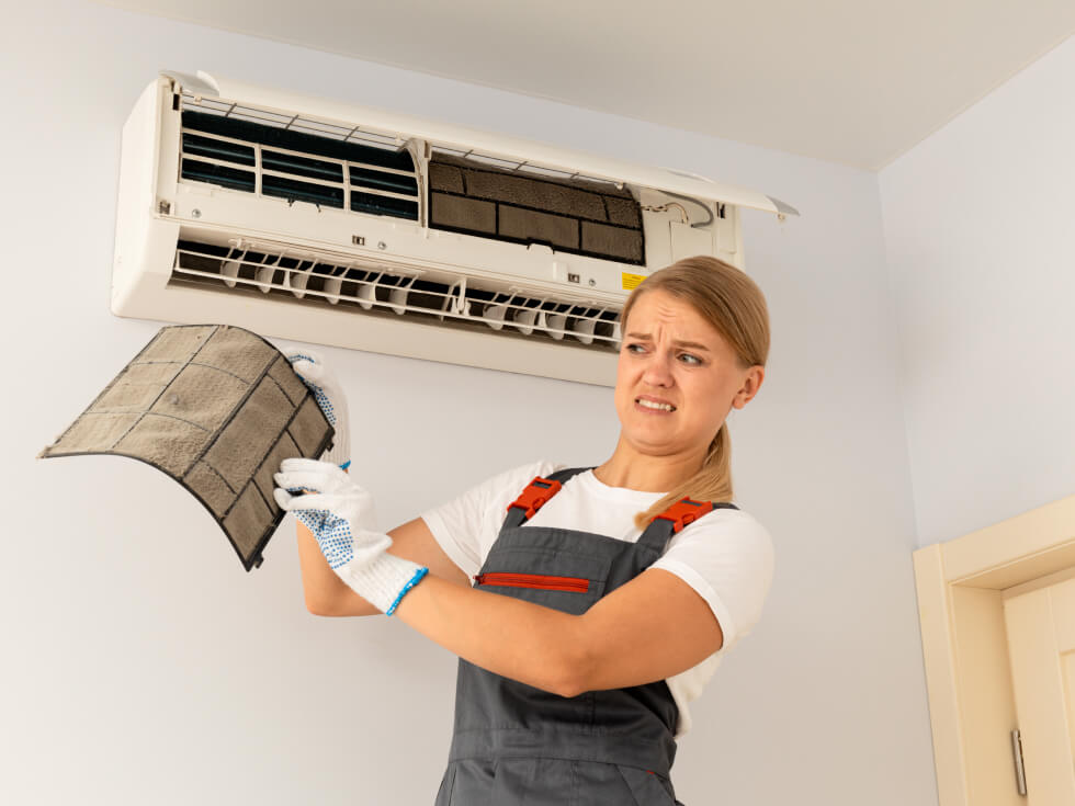 A woman wearing gloves looks at a dirty air conditioning filter they removed from the wall unit.
