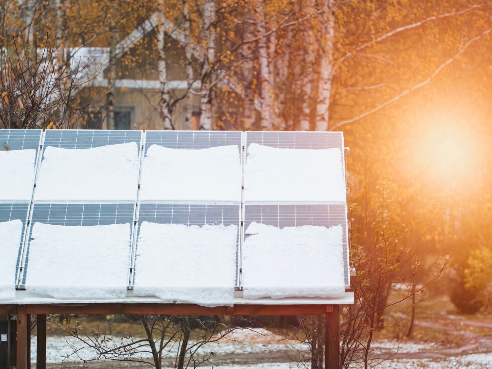 A solar panel partially covered in snow is shown in front of a house with autumn trees in the background.