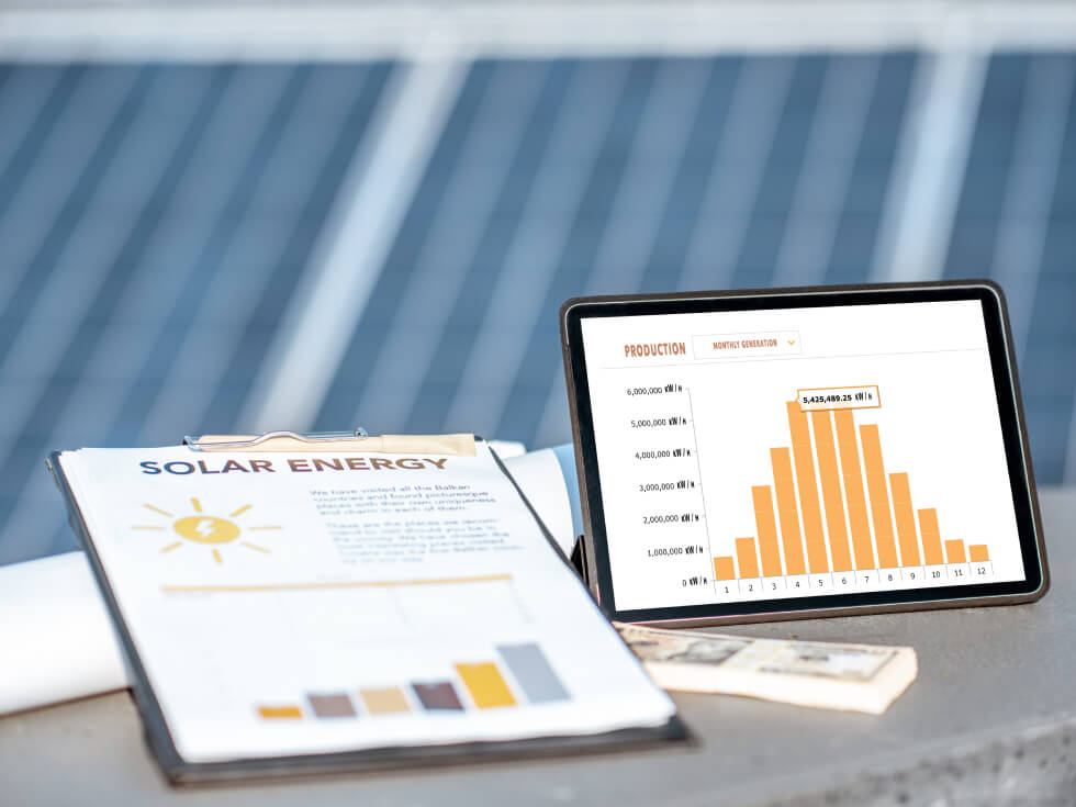 A tablet displaying a bar chart titled "Production" is placed next to a clipboard with a "Solar Energy" document in front of solar panels.