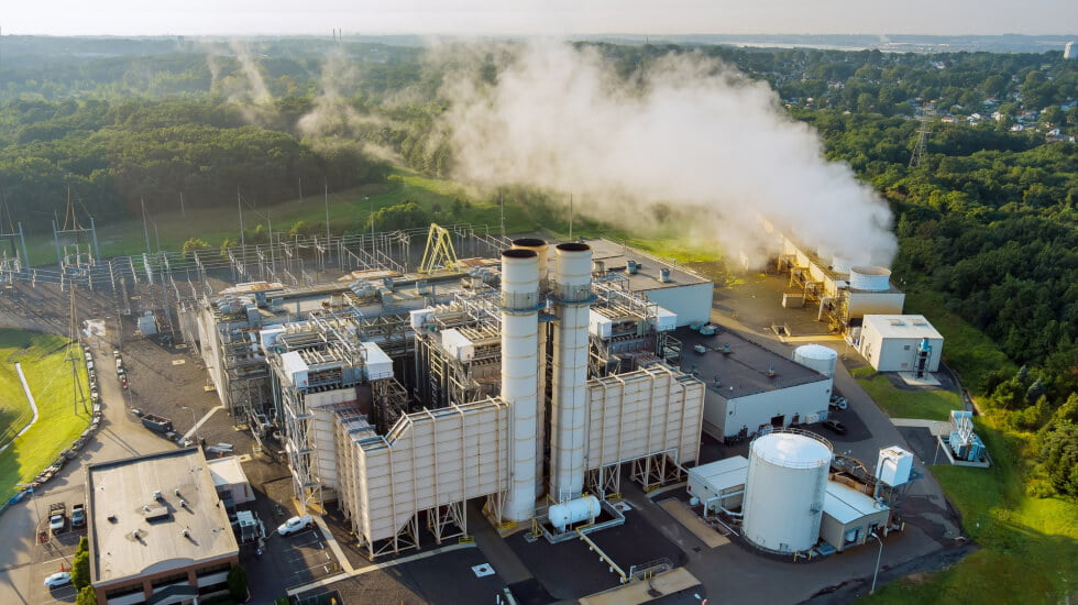 Aerial view of an biomass powerplant facility with two large smokestacks emitting white smoke, surrounded by greenery and buildings.