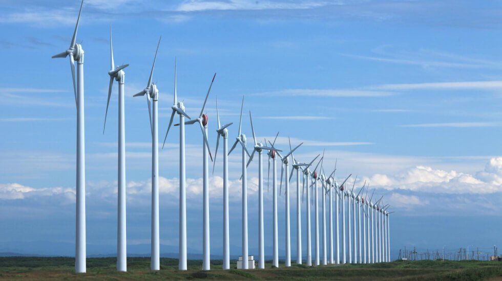 A row of large white wind turbines in an open field under a blue sky with scattered clouds.