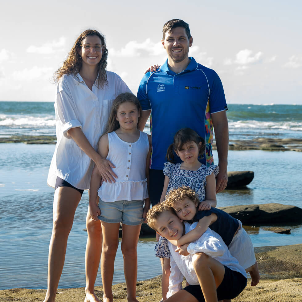 Tenmen Electrical family of five poses on a rocky shore on the Sunshine Coast, with parents standing behind and four children in front, against a backdrop of water and sky.