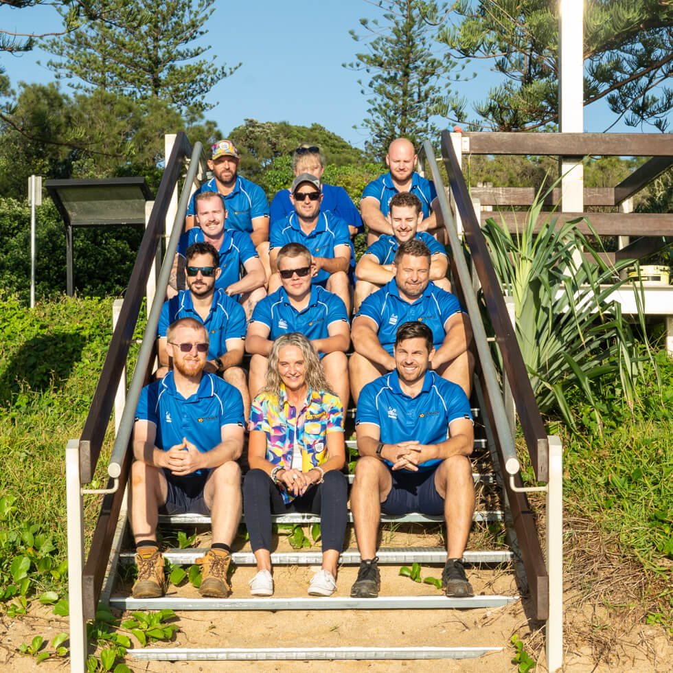 Tenmen Electrical team wearing the blue uniform sitting on outdoor stairs in a green, wooded area, with one person in a colorful shirt in the front row, also sitting.